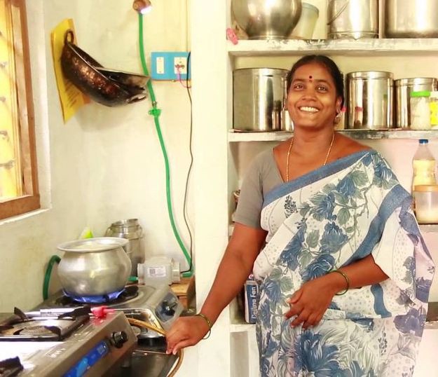 A woman smiling at a kitchen stove, a biogas line running down the wall to the burner