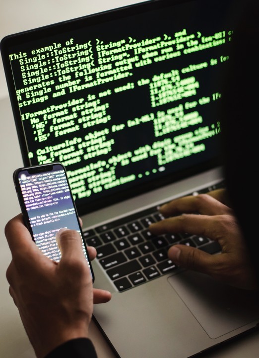 A person holding a phone in front of a laptop whose screen is filled with green text on black