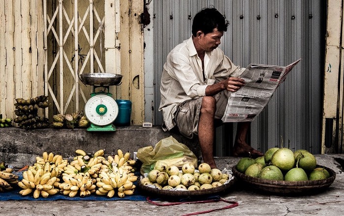 A market vendor reading a newspaper beside a mechanical dial weighing scale, with bananas, limes and coconuts laid out in front of him